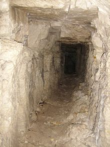 A narrow first World War fighting tunnel carved through chalk is depicted. The tunnel, roughly the height of a person, is illuminated by the white of the chalk, casting shadows along its walls. On the floor, there is rubble pilled to one side. The rough-hewn walls bear marks of manual excavation.