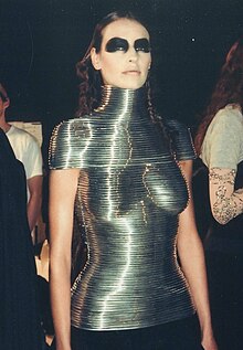 A woman wearing a form-fitting corset made of coiled metal, which covers her from the neck to the hips, and from the shoulder to the upper arm. She wears a black skirt and heavy black eye makeup.