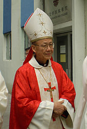 A bespectacled Chinese man wearing a white and gold-coloured mitre, red vestments and a pectoral cross faces towards the right.