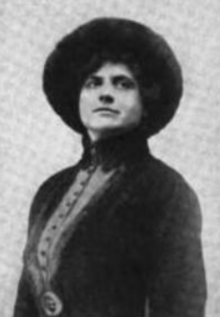 A white woman wearing a dark hat and a matching fitted jacket with a high collar.