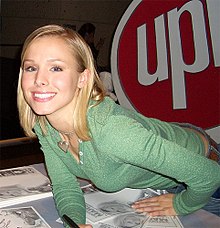 A blond woman wearing a green top leans forward over the table to smile at the camera to her left