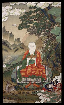 Elderly monk in green and red robes, holding his hands in front of his chest.