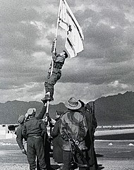 Israeli soldiers stabilize a flag pole whilst another soldier climbs it in order to raise an improvised flag; the soldier is seen about halfway up the flag pole. Other soldiers look on.