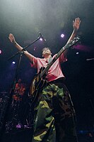 An image of Clinton Kane from a below point-of-view. He is wearing green camo cargo pants and a reddish pink short-sleeve button-up shirt. He has a guitar hung on his shoulder, and is raising his hands out forward and above his head.