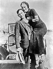 A sepia photograph, showing a man and a woman in front of a vehicle.
