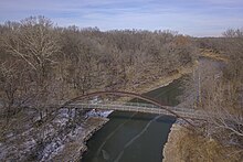 An aerial image of the Pathfinder Parkway bridge, a suspension footbridge with wooden planks, and the Candy River, with spots of snow on the ground