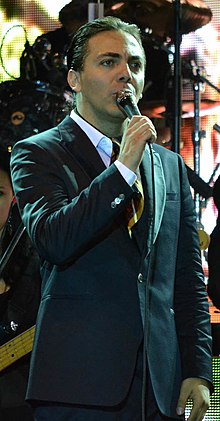 Man with black-dyed hair is wearing a tuxedo and holding a microphone on his right hand
