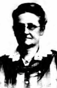 An older white woman, wearing eyeglasses and a lace collared dress