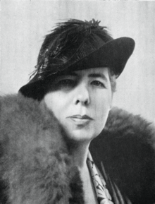 Black and white portrait photograph of Gordon Holmes. Framed so we can only see her from the shoulders upwards, she is turning to look into the camera - a middle-aged woman wearing a black hat, with a large fur draped around her shoulders, and bearing a resolute expression on her face.
