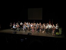 Fiddle players holding their fiddles gather on a stage in front of an audience