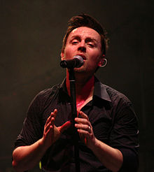Upper body photo of a thirty-four year-old man. He is shown facing forward and singing into a microphone with eyes partly closed. Both hand are raised in front to chest level. He wears a dark coloured, short-sleeved shirt. He wears a ring on his left fourth finger.