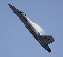 Jet fighter aircraft is seen against blue sky executing a pull-up, making it nearly vertical with contrail formed aft of the canopy