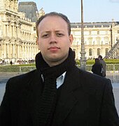 Tenor Ewandro Stenzowski in front of the Louvre museum in Paris, France. He is wearing a black coat and black scarf over a white button down shirt.