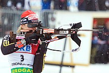 A woman in multicoloured winter sportswear, wearing a red cap and a jersey with the number 3, holds a rifle in a horizontal position. Her rifle has advertising on its side, while snow falls in the background.