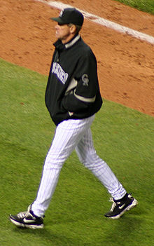 A man in white baseball pants, a black jacket, and black cap