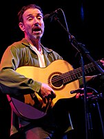 A man with a goatee singing into a microphone playing a guitar.