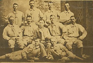 A baseball team is posing for a photograph. There are four men standing, five men sitting, and two men are laying on the ground.
