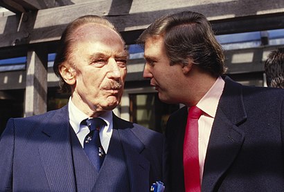 On the left, a decrepit old man with an unusual jawbone deformation underlying his mildly baggy skin. He wears a blue lined suit and vest with a blue tie and puckers an expression of interest at the younger man at his right, who leans forward as if offering a cunning suggestion. The youth, seen in profile, wears a red tie and woollike coat with a lined shirt underneath. Behind them is a passerby, above them rafters.