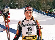 A young woman in multicoloured winter sportswear and with the number 19 on her jersey smiles into the camera, standing in a field covered with snow. A forest and many onlookers can be seen in the background. She holds ski poles in her right hand and has a rifle on her back.