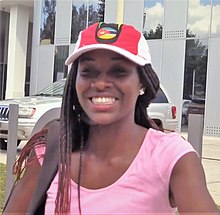 Headshot of a black woman wearing a baseball cap with a Mozambique flag on it, with a glass building and white car behind her