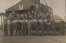 In a black and white photograph, group of 26 young to middle-aged men wearing suits and flat caps stand in two rows. Behind them is a large, industrial shed, and in the distance the roof of a house can be seen.