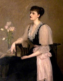 Portrait of a pensive Violet Jacob, wearing a pink and black gown with ruffle at the neck, seated beside a vase of flowers. Oil on canvas.