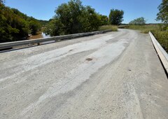 A short and unmarked concrete road bridge, with white gravel scattered atop, spans a brown creek with trees and grasslands on either side; graffito on the visible guardrail says, "GONE NOT FORGOTTEN ♥ RIP HEATHER 1996 ♥"