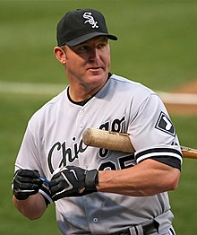 A man in a grey baseball uniform with "Chicago" on his chest in black with a black cap, black batting gloves, and a baseball bat under his arm.