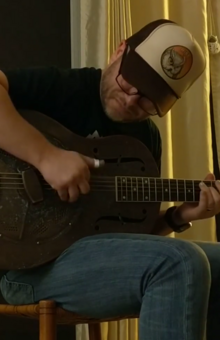 Ross Hammond is playing a resonator guitar. He is seated on a chair, bent over the guitar, wearing a trucker hat and glasses.