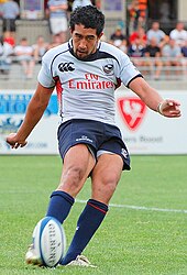 A man wearing a rugby uniform consisting of a white shirt, dark blue shorts, and dark blue socks attempts a drop kick.