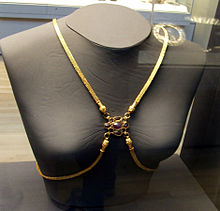Frontal view of a grey female bust with four gold bands of many fine links, two draped over the shoulders and two plunging from the breast bone sweeping below the breasts and going behind the back. The four converge between the breasts, where each band ends in a head which connects to a centerpiece, a purple gem surrounded by eight smaller sockets, four empty and four with red stones
