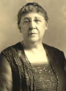 Annie Laws in later life, a white woman with grey hair, wearing glasses and a dark dress with a square neckline and sequined or beaded details