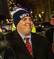 A front-facing photograph shows at bust-length standing and smiling. His head is looking to his left. This photograph shows Flynn is outdoors at night in a well-lit area. He is wearing a blue puff beanie cap with the word "Navy" printed above its rim, the hat is pulled down to nearly cover the entirety of his ears. He is also wearing a formal jacket, with a blue shirt and a necktie underneath. Other people are partially-visible standing in his proximity.