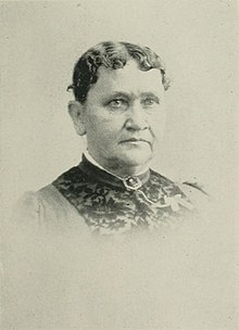 B&W portrait photo of a woman with her wavy hair in an updo, wearing a dark high-collared blouse.