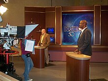 Two men stand at lecterns on a set with KVIE logos in front of a camera