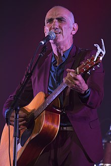 Head and shoulders of a sixty-year-old male standing at a microphone. He is strumming a guitar, singing and staring into the distance. He wears a purple suit, tie, blue shirt and vest.