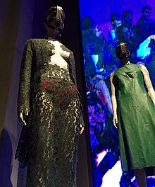 Two mannequins seen from a low angle. The left mannequin wears an outfit made from black lace, with the centre of the chest exposed and red underwear visible underneath on the pelvis. The right mannequin wears a sleeveless green leather sheath dress with an open slash over the breast area.