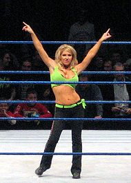 A blond woman poses in a wrestling ring with blue ropes. Her arms are outstretched, and she is pointing to either side. She is wearing a green crop top, with a matching green belt, and black trousers.
