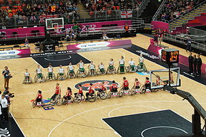 Parallel lines of two teams of players in wheelchairs, one in red, the other in green and white. They are on a basketball court, surrounded by media, official in black, and spectators.