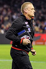 A man with light brown hair in a black jacket jogs across a grassed playing field