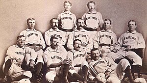 In this photograph of a baseball team, eleven men are situated in three rows facing the camera, with four sitting on the floor, five sitting in chairs, and two standing.