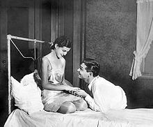 A young woman in a slip dress is kneeling on a bed while smiling at the young man clasping her hands, who is laying in a prone position in a dress shirt and pants and is smiling back.