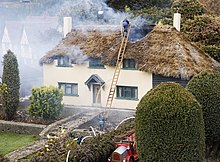 The roof of a thatched miniature house smokes as firemen attend