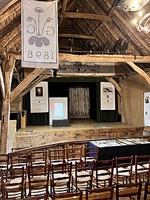 The interior of a large wooden barn, which has been converted to a theatre, with a view of the stage and two rows of wooden chairs