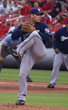 A man wearing a navy blue Brewers jersey, gray pants, and a navy blue cap shown preparing to throw a ball from the pitcher's mound