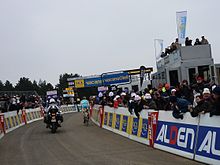 Many spectators in warm clothes stand behind barricades watching a lone cyclist in a blue and yellow jersey with white trim. A motorcycle follows the cyclist.