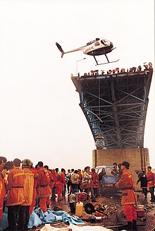 View of rescue operations from on the collapsed section of the bridge. First responders are wearing orange uniforms, and a helicopter is visible flying above the bridge.