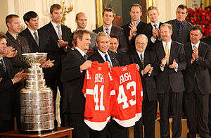 In the front row, Nicklas Lidstrom holds a Red Wings jersey with the last name "BUSH" and the number "41" while George W. Bush holds a Red Wings jersey with the last name "BUSH" and the number "43". To Lidstrom's right is the Stanley Cup. Second row, left to right: Mike Babcock, Ken Holland, Jim Devellano, Jim Nill, unknown man. Third row, left to right: Kris Draper, Pavel Datsyuk, Mikael Samuelsson, Brian Rafalski, Derek Meech, Kirk Maltby, Chris Osgood, Aaron Downey.