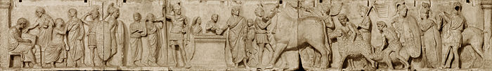 Relief depicts a long continuous scene that may be divided into three main groupings from left. The first is a group of four men wearing togas. Two are seated, and one is writing in a tablet. Two tall military guards divide this group from the central scene of sacrifice. Two musicians, markedly shorter than the soldiers, play a lyre and a horn. An unadorned altar, waist-high, stands in the center. To the viewer's left is the tallest figure in the composition, a military officer wearing a high plumed helmet and holding a long slender spear. standing by an altar. On the other side of the altar a priest, his head ritually covered, extends a libation bowl. A boy attendant pours from a pitcher into the bowl, and to that boy's right is a smaller boy looking on and lifting his right hand to the top of his head, a gesture that appears quizzical to modern viewers but may have some other significance in its Roman context. The priest is accompanied by a third boy close to his left side who stands ready with a towel. The right side of the relief is devoted to the procession of the three animal victims for the suovetaurilia, each led by a young male attendant, barechested but wearing a short kilt-like garment, with a wreath on his head. The first leads an enormous bull with a tasseled rope dangling from below its left horn. A fourth male attendant in the same attire follows closely on the bull's hindquarter, waving a palm branch in each hand. The attendant bringing the ram is followed by another veiled figure carrying a pole from which a banner unfurls. The attendant herding the pig is followed by another soldier bearing a long shield and looking back at another whose shield rests on the ground, covering most of his body. The last figure is a cavalryman, back turned to the viewer, next to his horse.