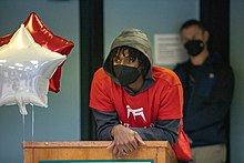 Maxey, wearing a red t-shirt over a gray hoodie and in a mask, leans over a podium.